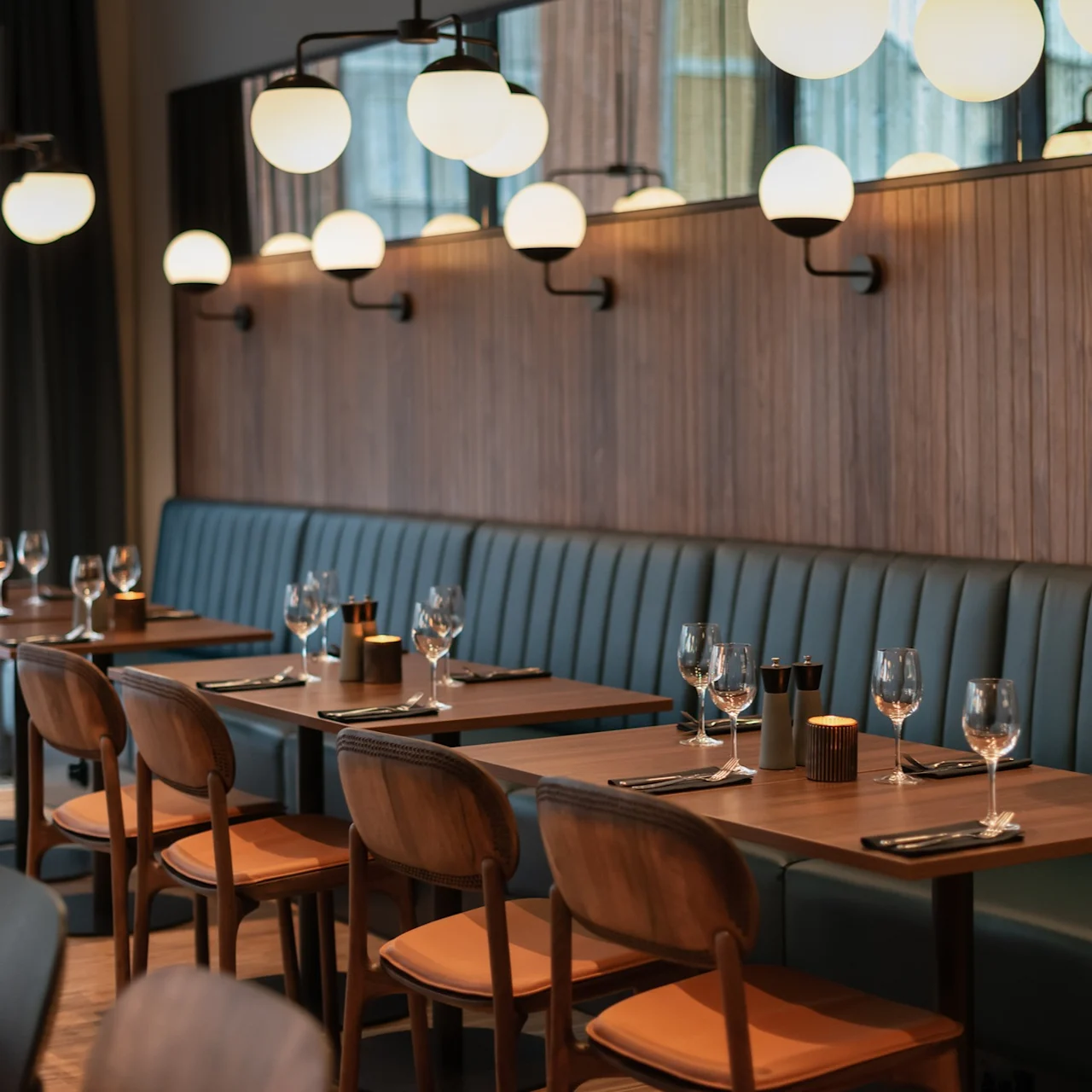 Bord dekket med glass og bestikk er arrangert i et stilrent, moderne restaurantinteriør med trevegger og myk belysning fra sfæriske lamper ovenfor.