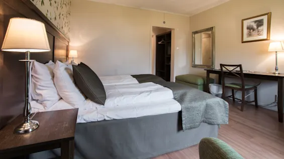 En snyggt bäddad säng med kuddar, inramad av sängbord och lampor, i ett välbelyst hotellrum med skrivbord, stol och spegel.