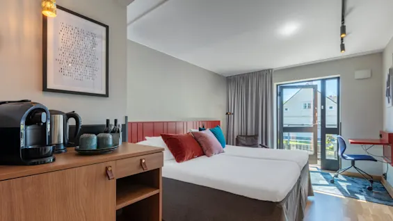 An inviting hotel room with a neatly made bed, wooden furniture, a coffee maker, and a framed text artwork, bathed in natural light from a balcony door.