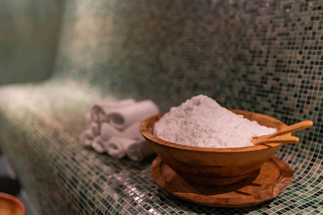 Puinen kylpysuolakulho kauhalla ja rullatuilla pyyhkeillä laatoitetulla pinnalla, luoden rentouttavan kylpyläkokemuksen.