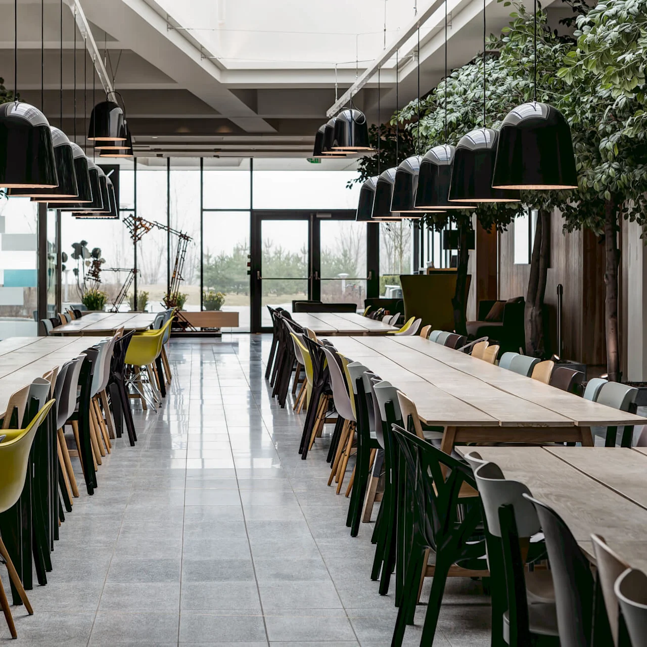 Long wooden tables with mismatched chairs are arranged under pendant lights in a modern cafeteria, flanked by indoor greenery.