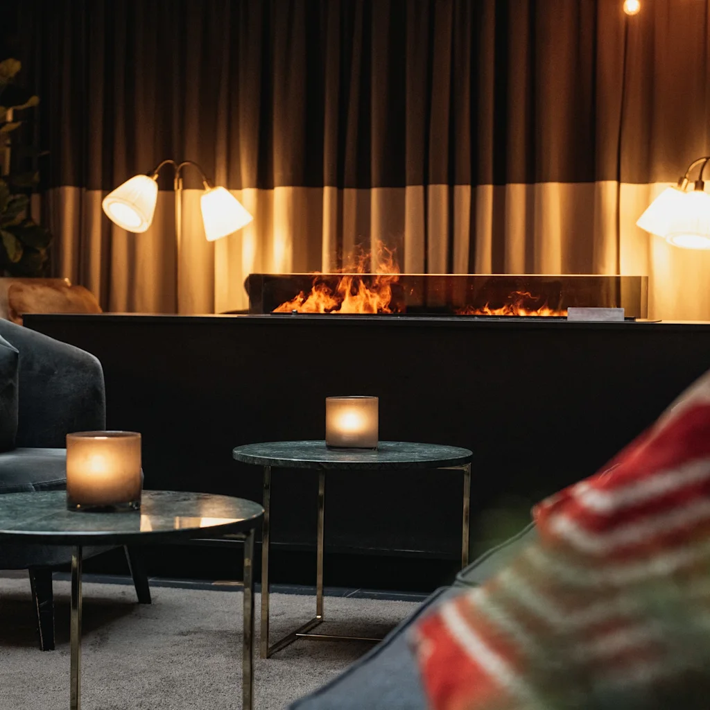 A cozy lounge area with a long fireplace, casting a warm glow. Candles on small tables add to the inviting ambiance.