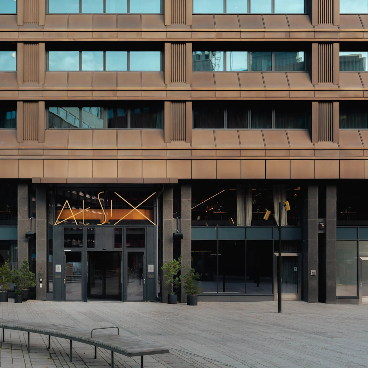 En brun fleretasjes bygningfasade med store vinduer og en moderne inngang med teksten "At Six." Pottede planter og en buet benk er plassert i gårdsplassen.