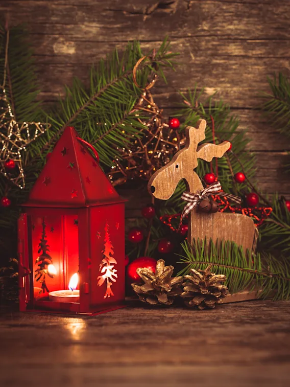Cozy Christmas decoration with a lit red lantern, a wooden reindeer figure, and pine branches on a rustic wood background.