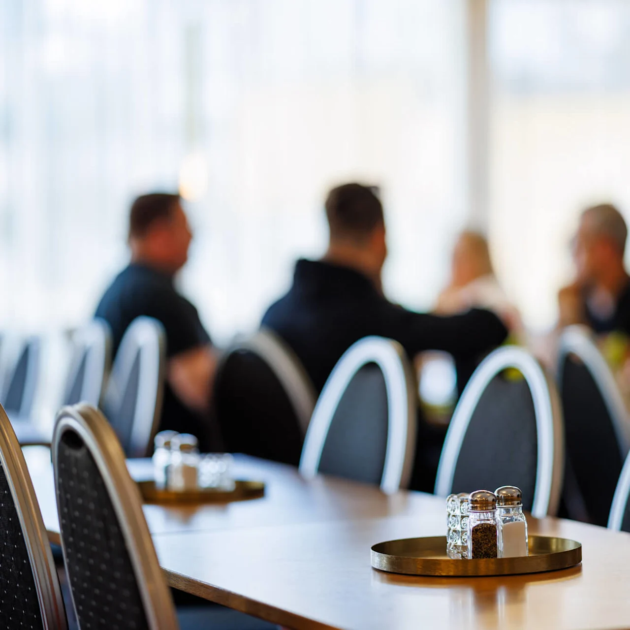En salt- och pepparströare står på en rund bricka på ett träbord i en restaurang; suddiga personer samtalar i bakgrunden.