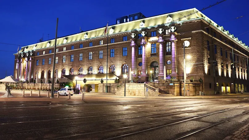 A large, illuminated hotel building with classical architecture stands against a dark blue evening sky. Several columns and windows are visible. "Clarion Hotel Post" signage is lit above the entrance.