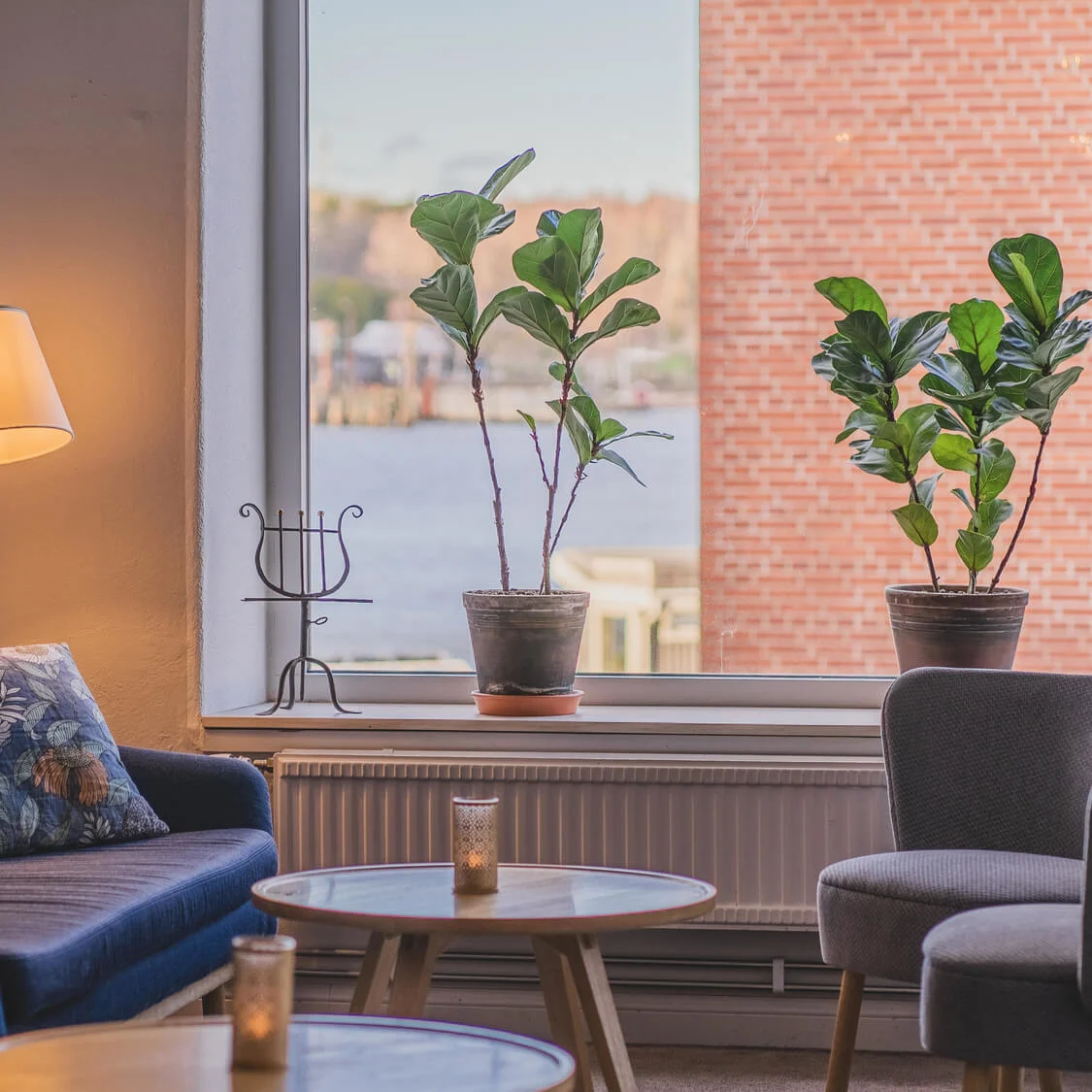 A cozy living room features a blue sofa with patterned cushions, a lit floor lamp, and round coffee tables. Large window showcases potted plants with a waterfront view.