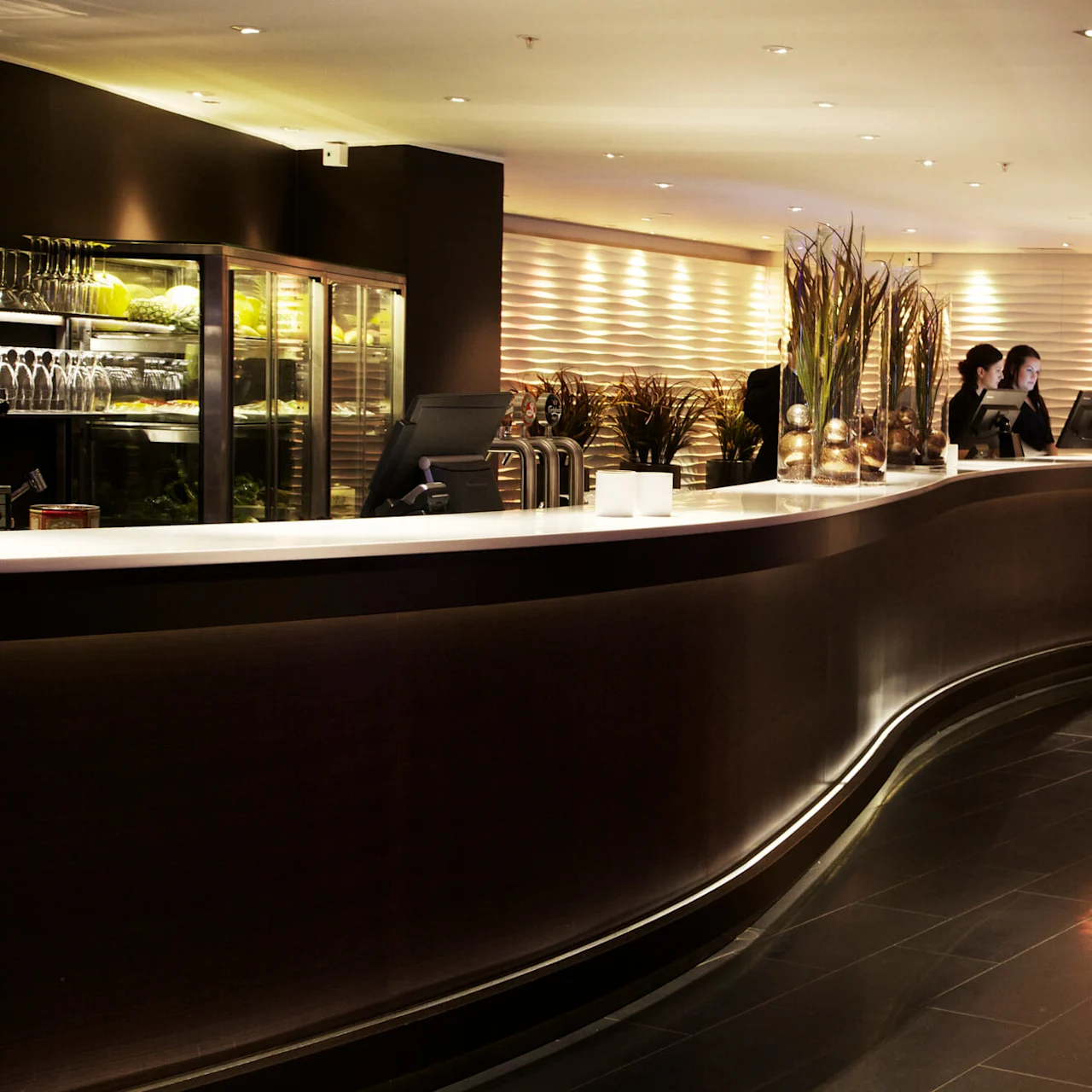 En böjd receptionsdisk med två personer som arbetar vid datorer, omgiven av dekorativa växter och stämningsljus, i en modern, varmt upplyst lobby.