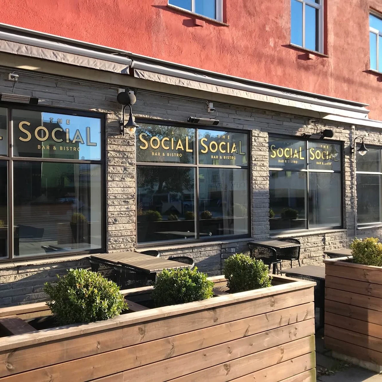 Norwegian (bokmål): Svartkantede vinduer viser "THE SOCIAL BAR & BISTRO" i gullbokstaver. Uteservering og treplanter med busker kantar steinfasaden.