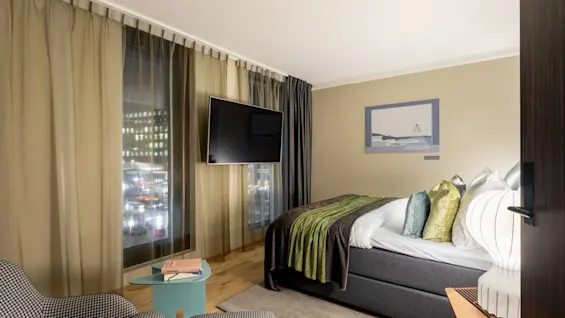 A bed with green and white bedding stands in a modern hotel room. A wall-mounted TV faces the bed, and large windows reveal city lights outside. Chairs and a table complete the space.