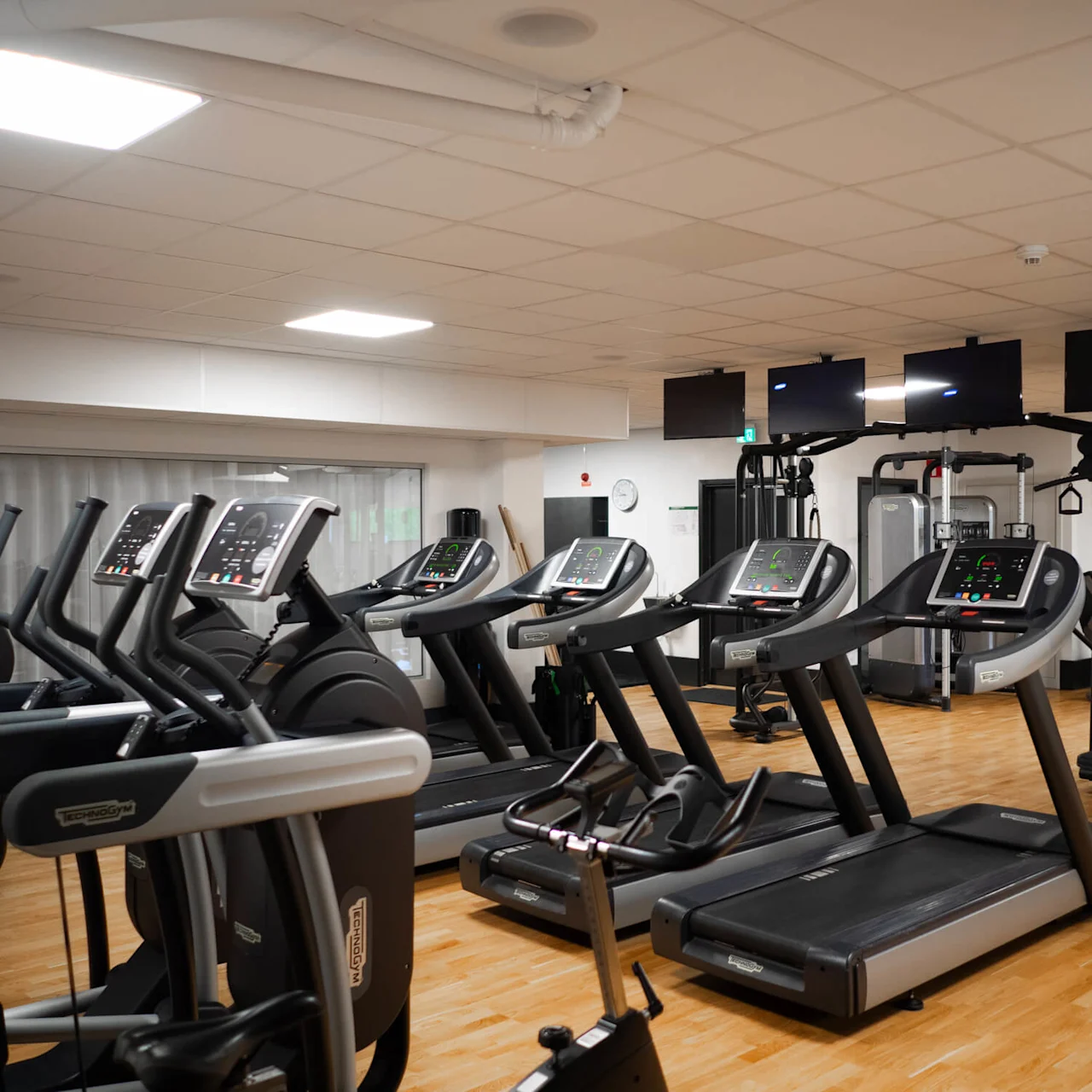 Löpband uppradade i ett ljust, rymligt gym med trägolv och träningsmaskiner. Väggmonterade tv-apparater ovanför löpbanden som erbjuder underhållning för användare.