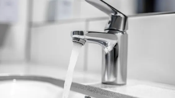 A chrome faucet flows water into a white sink, surrounded by white tile walls and a soap dispenser in a bathroom setting.