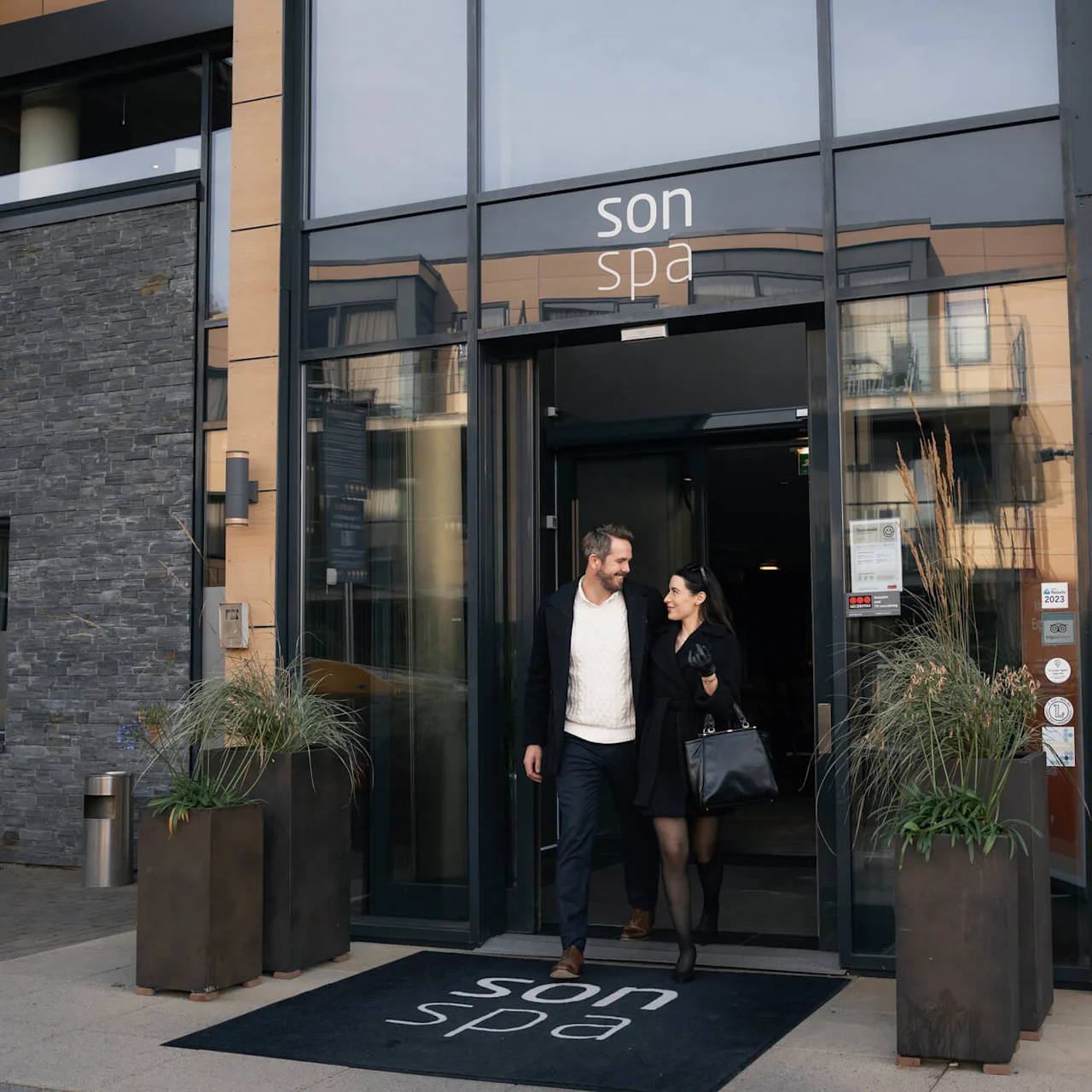 Et par kommer ut gjennom glassdørene til en bygning merket "son spa", med planter på hver side, og reflekterer et moderne, urbant miljø.