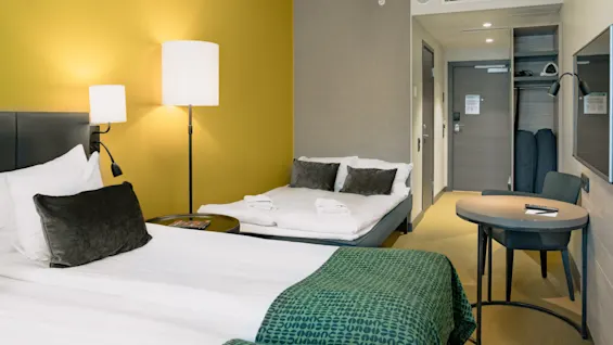 Ett prydligt ordnat hotellrum med två sängar med vita lakan, åtföljt av ett runt bord och stol, upplyst av stående lampor mot senapsfärgade och grå väggar.