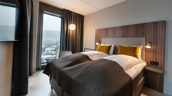 En pent, dobbeltseng med brunt og hvitt sengetøy står mot en trefinérs hodegjerde. To lamper gir et varmt lys. Store vinduer med mørke gardiner viser en byutsikt.