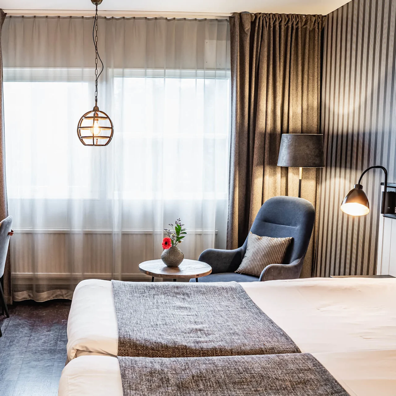 A neatly arranged hotel room features a bed, desk with lamp and chair, armchair with pillow beside a window, and a hanging light. Curtains and striped wallpaper add elegance.