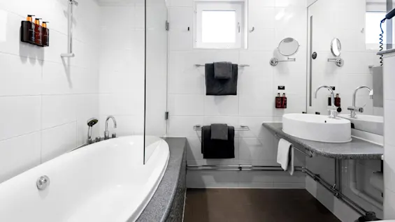 A modern bathroom with a white bathtub, shower, sink, and towels. Reflective surfaces and a window for light.