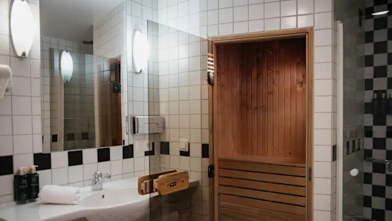 Bastu med träinteriör intill ett badrumshandfat. De vitkaklade väggarna, den stora spegeln och belysningen skapar en ren, ljus miljö.