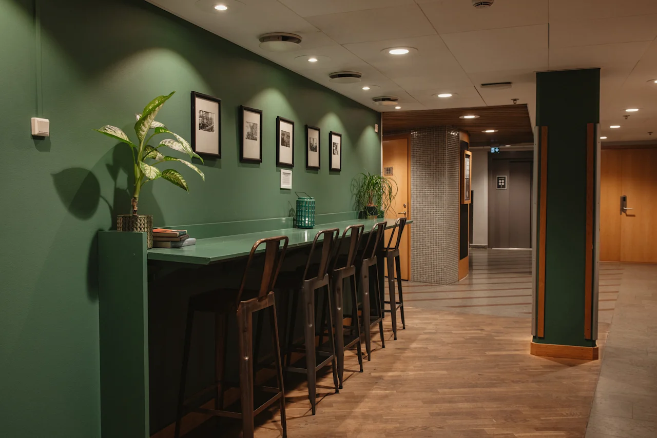 A modern hotel lobby with a long green counter and bar stools, framed art on the wall, and plants. Home Hotel Uppsala.