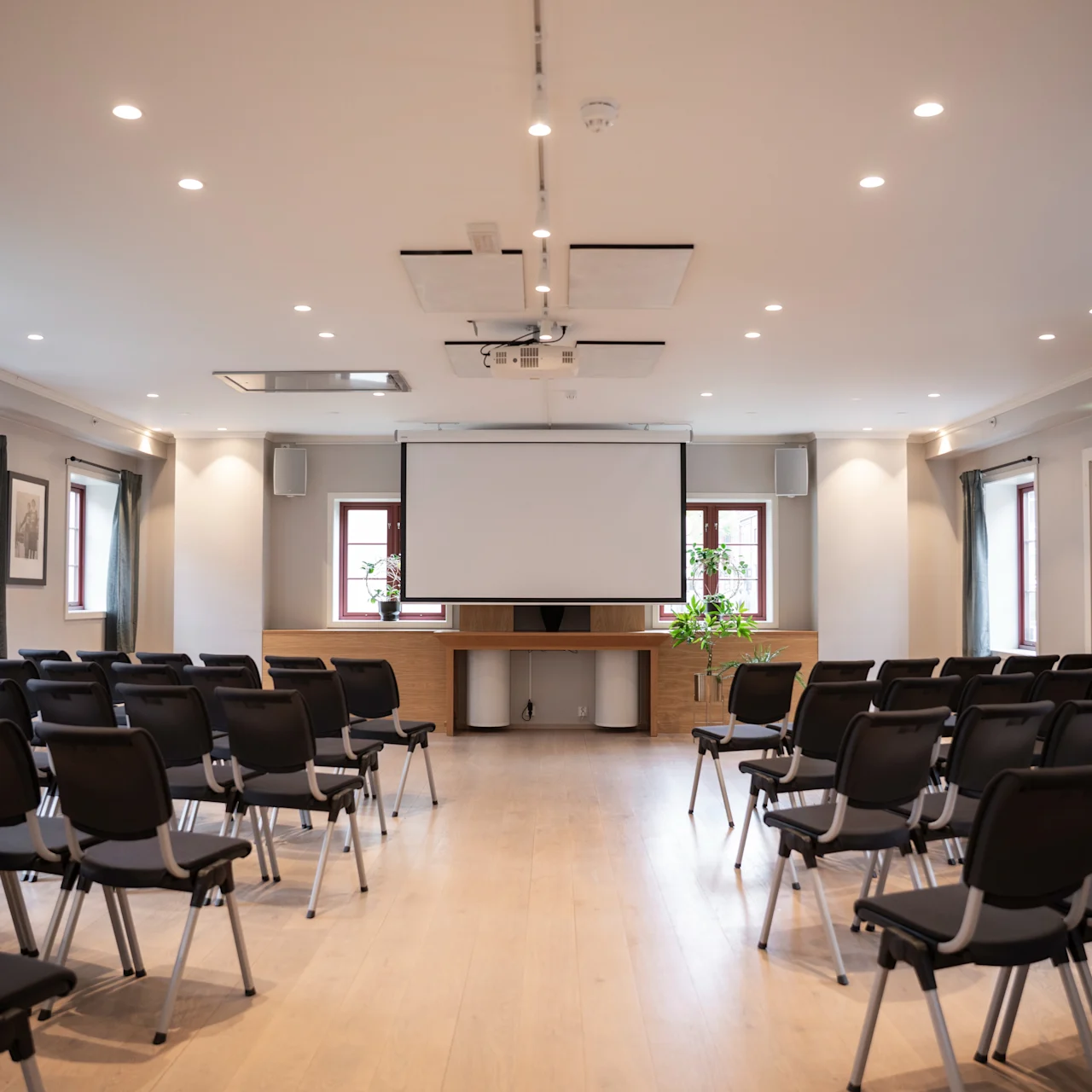 Konferensrum med rader av tomma stolar vända mot en stor projektorduk. Rummet är väl upplyst med taklampor och har inramade bilder och fönster i en modern miljö.