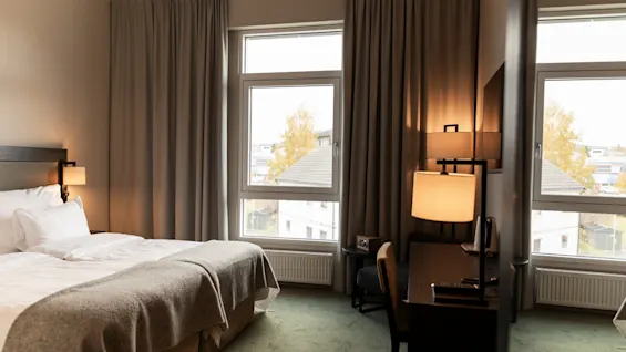 Et hyggeligt hotelværelse med to senge, et skrivebord og store vinduer med udsigt til omgivelserne.