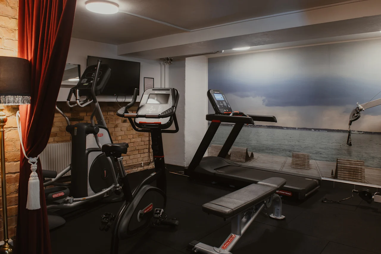 Ett välutrustat gym på Home Hotel Grand, Helsingborg, med olika träningsmaskiner och en naturskön väggmålning av en strandpromenad.