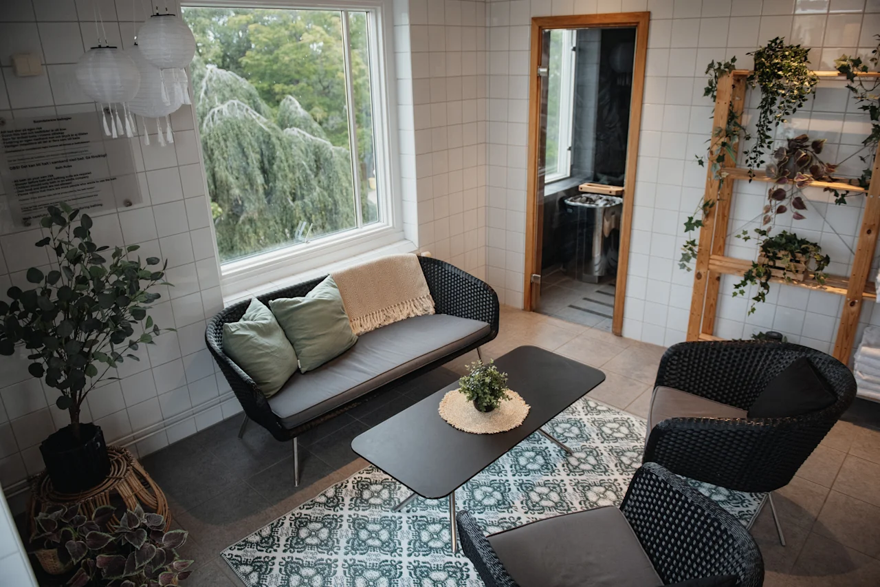 Relaxation area with a view of trees, sofa, armchairs, and a coffee table. Sauna entrance. Sign: Regler for bruk av badstuen.