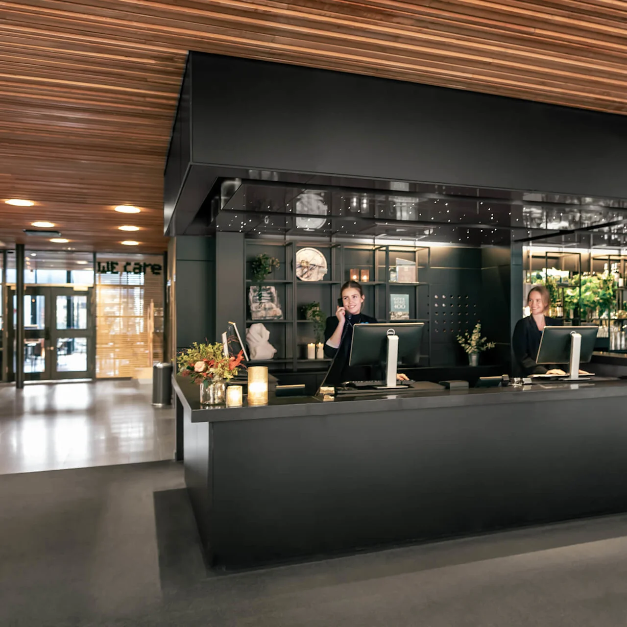Reception desk with two staff members assisting guests, set in a modern hotel lobby featuring a wooden ceiling, decorative plants, and ambient lighting. Text: "WE care".