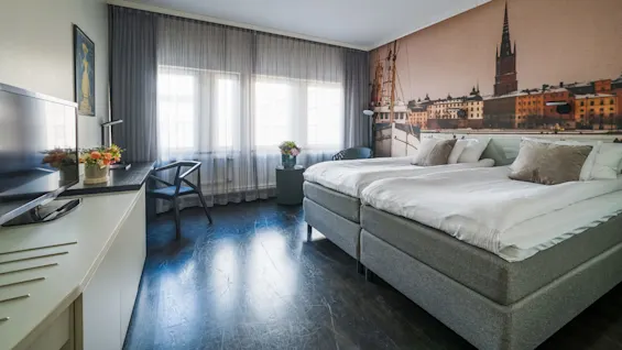 A neatly arranged hotel room features a large bed with pillows, a desk with flowers, a chair, and a TV facing large windows with sheer curtains, creating a bright ambiance.