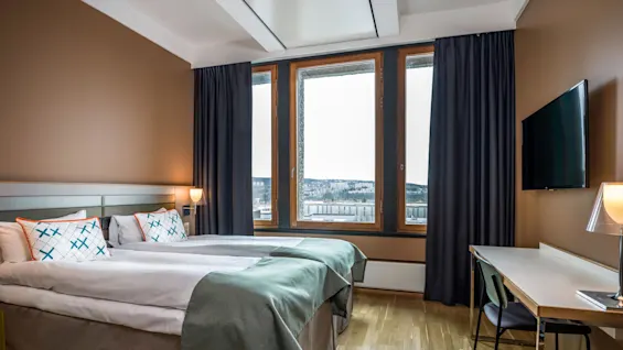 A tidy hotel room with a neatly made bed beside large windows offering a snowy landscape view, a desk with a chair, and a wall-mounted television.