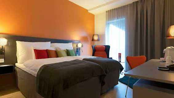 En prydligt bäddad säng med färgglada kuddar mot en orange vägg, flankerad av lampor. En stol och ett skrivbord med kaffemaskin finns nära skira gardiner.