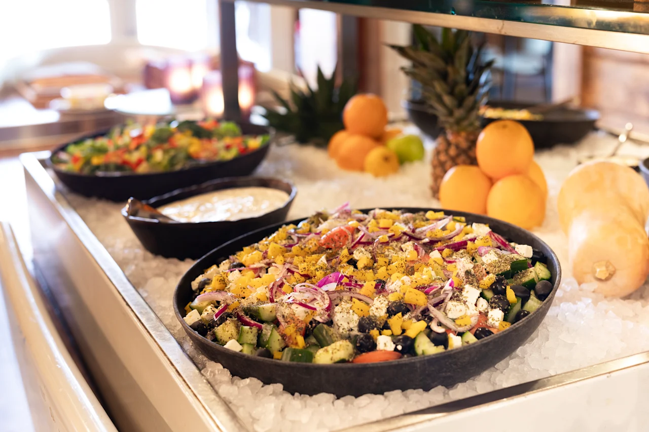 Et levende buffetbord med friske salater, frugter som ananas og appelsiner samt en cremet dressing, alt afkølet på is.