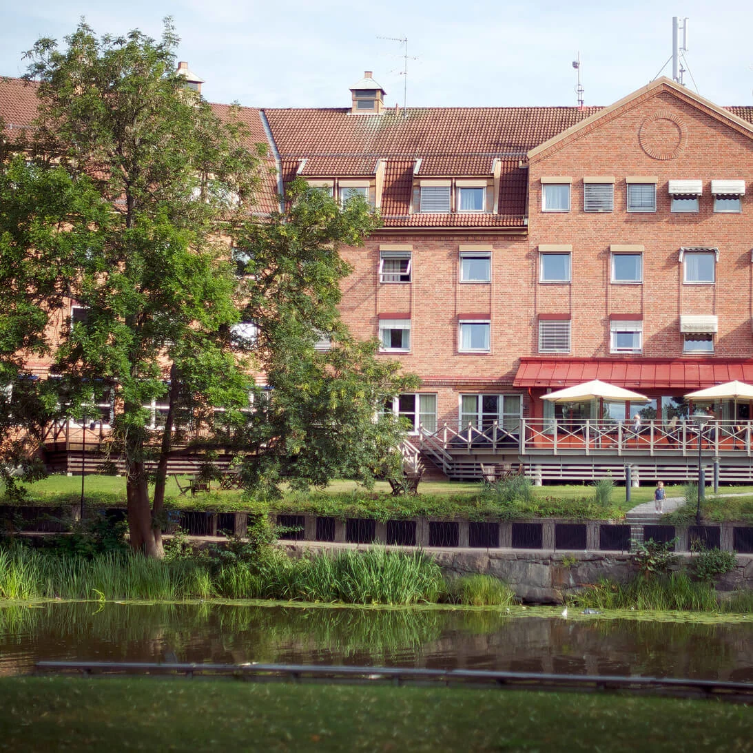 A red brick building with large windows and a terraced café overlooks a tranquil water body surrounded by greenery.