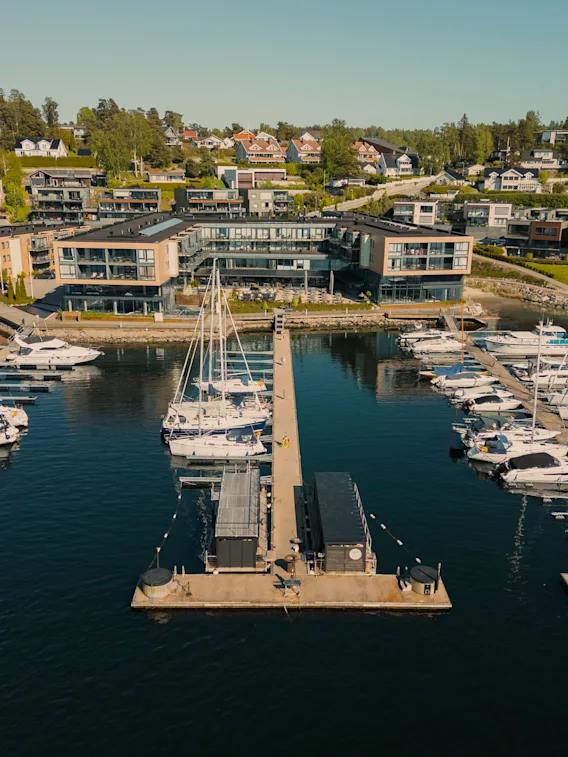 En flyutsikt over en livlig marina med mange båter fortøyd, mot et bakteppe av moderne bygninger og hus i åssiden.