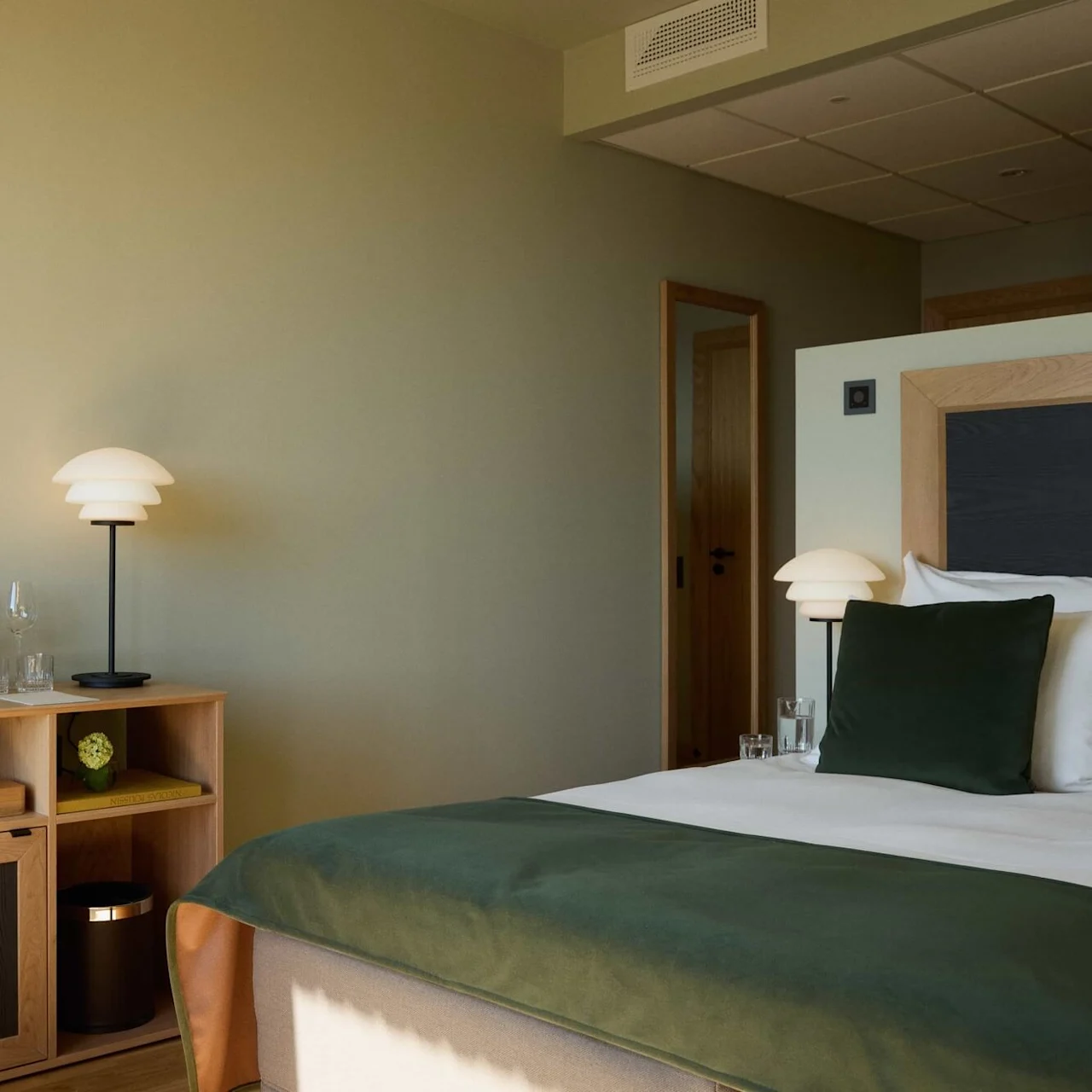 A neatly arranged hotel room features a bed with green accents, a wooden shelf with a lamp and a coffee machine. Neutral walls and warm lighting create a peaceful atmosphere.