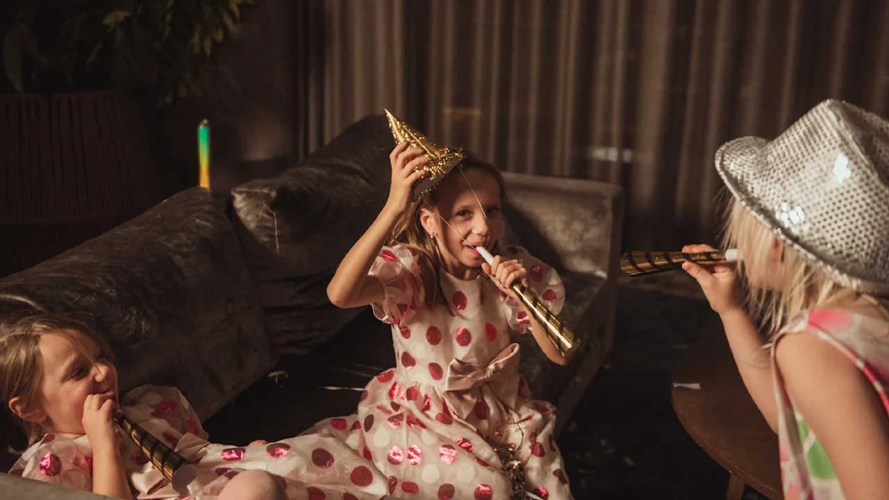Three children wearing festive hats blow party horns, sitting on a couch in a dimly lit room, celebrating together.