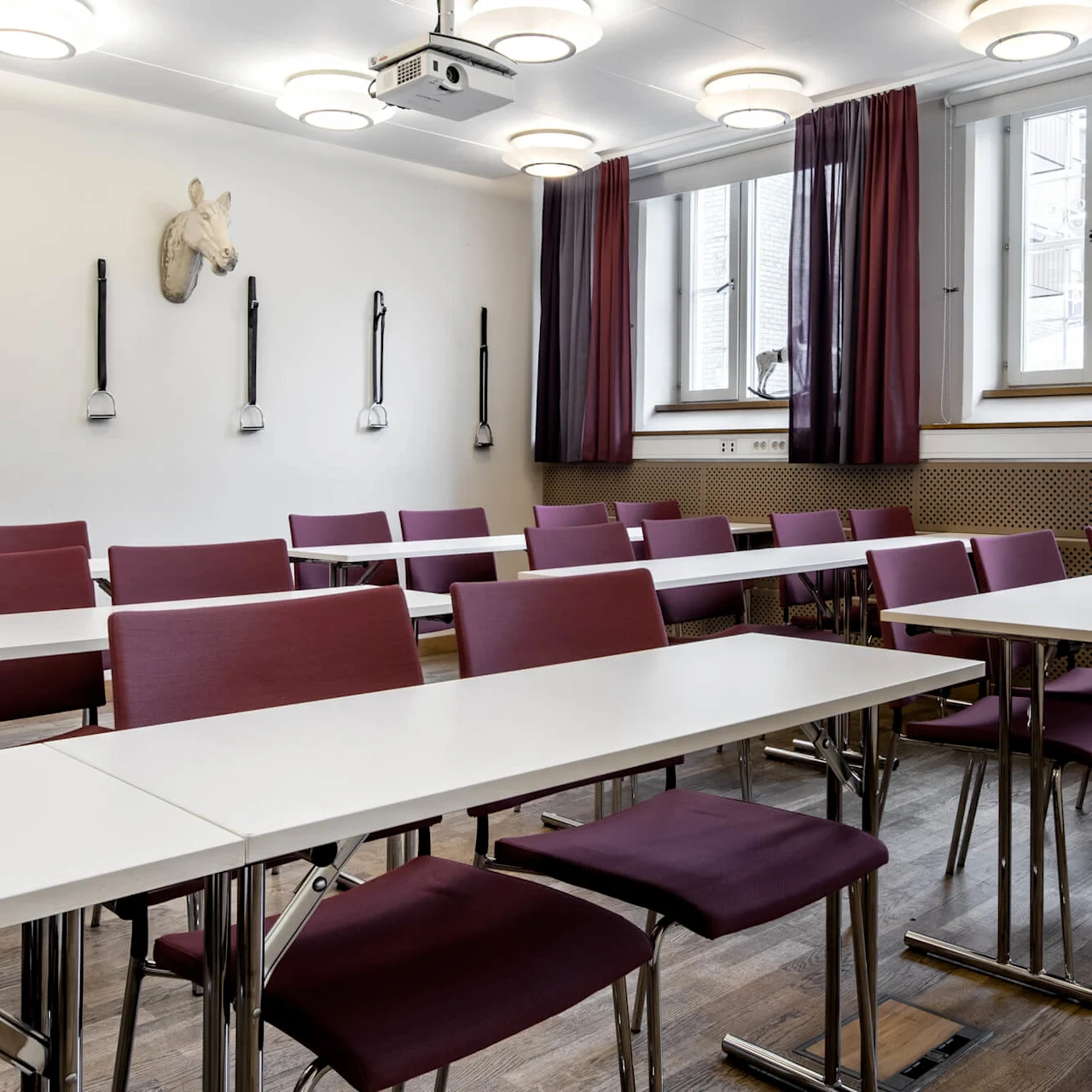 A conference room with rows of white tables and maroon chairs, illuminated by ceiling lights.