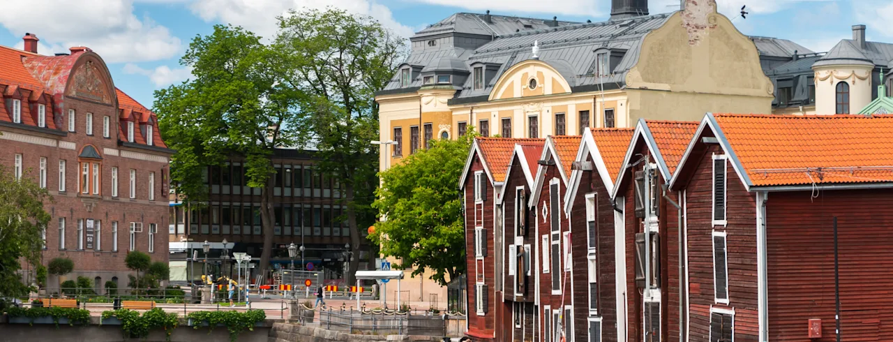 Röda trähus står vid en flodkant och speglas i lugnt vatten. Bakom dem finns historiska stenbyggnader och lummiga träd under en klarblå himmel med några få moln.
