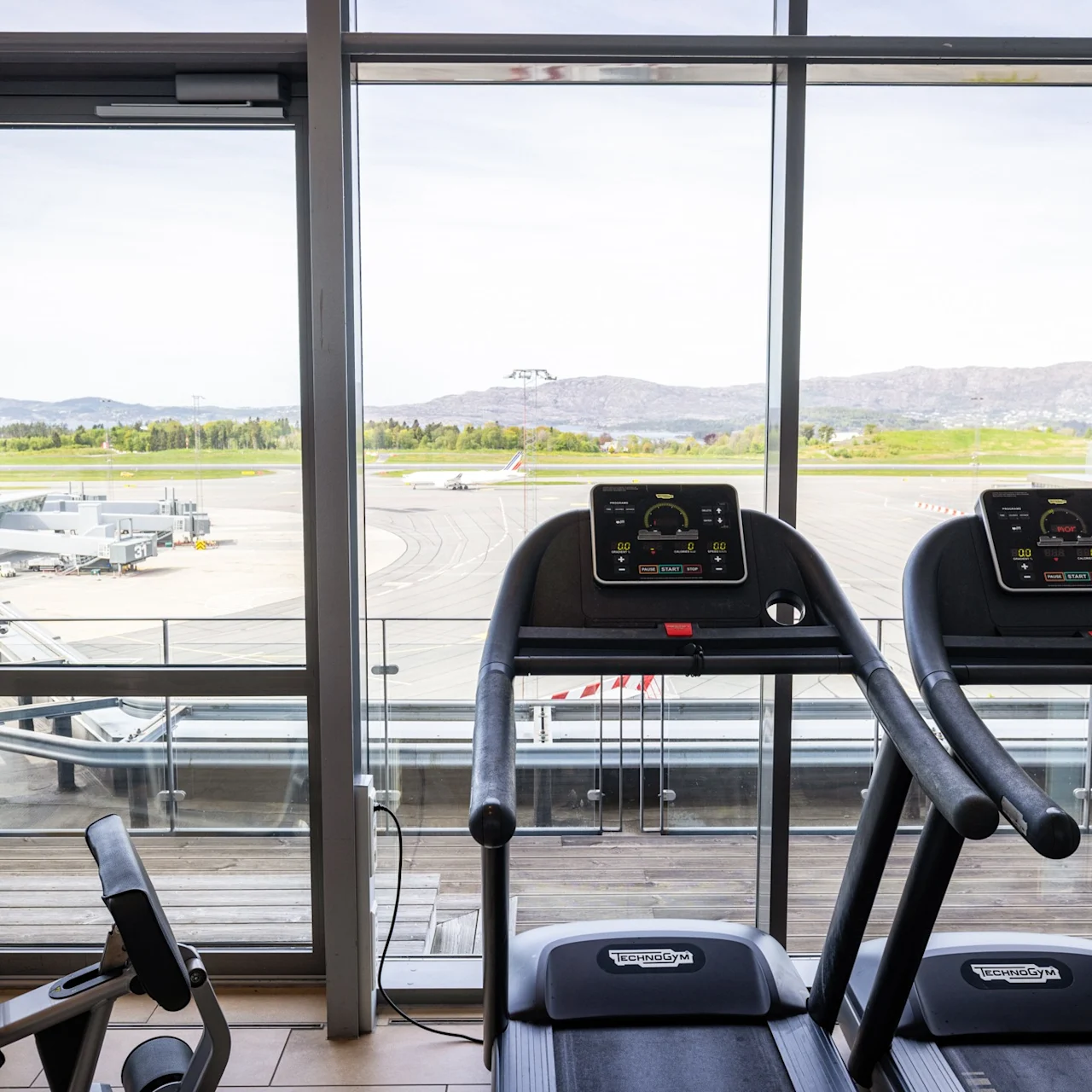 Treadmills positioned in front of large windows overlooking an airport runway with parked planes. Bright daylight illuminates the fitness area. No text visible.