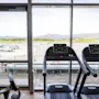 Tredemøller plassert foran store vinduer med utsikt over en flyplass med parkerte fly. Sterkt dagslys belyser treningsområdet. Ingen tekst synlig.