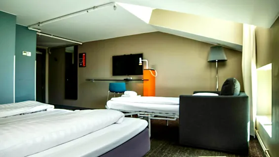 Ett hotellrum med flera sängar, en TV och en soffa, med snedtak och varm belysning.