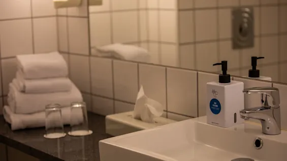 En hvit vask med kromkran er ved siden av en såpedispenser merket "GO CLEAN HAND SOAP" på et flislagt baderom. I nærheten er foldede håndklær, to glass og servietter.