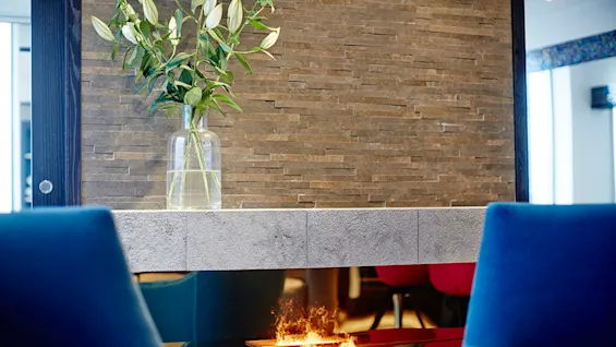 Glasvase med grønne blade står på en stenmantel. For neden flammer en moderne pejs. Blå stole indrammer forgrunden i et moderne rum med sten vægge.