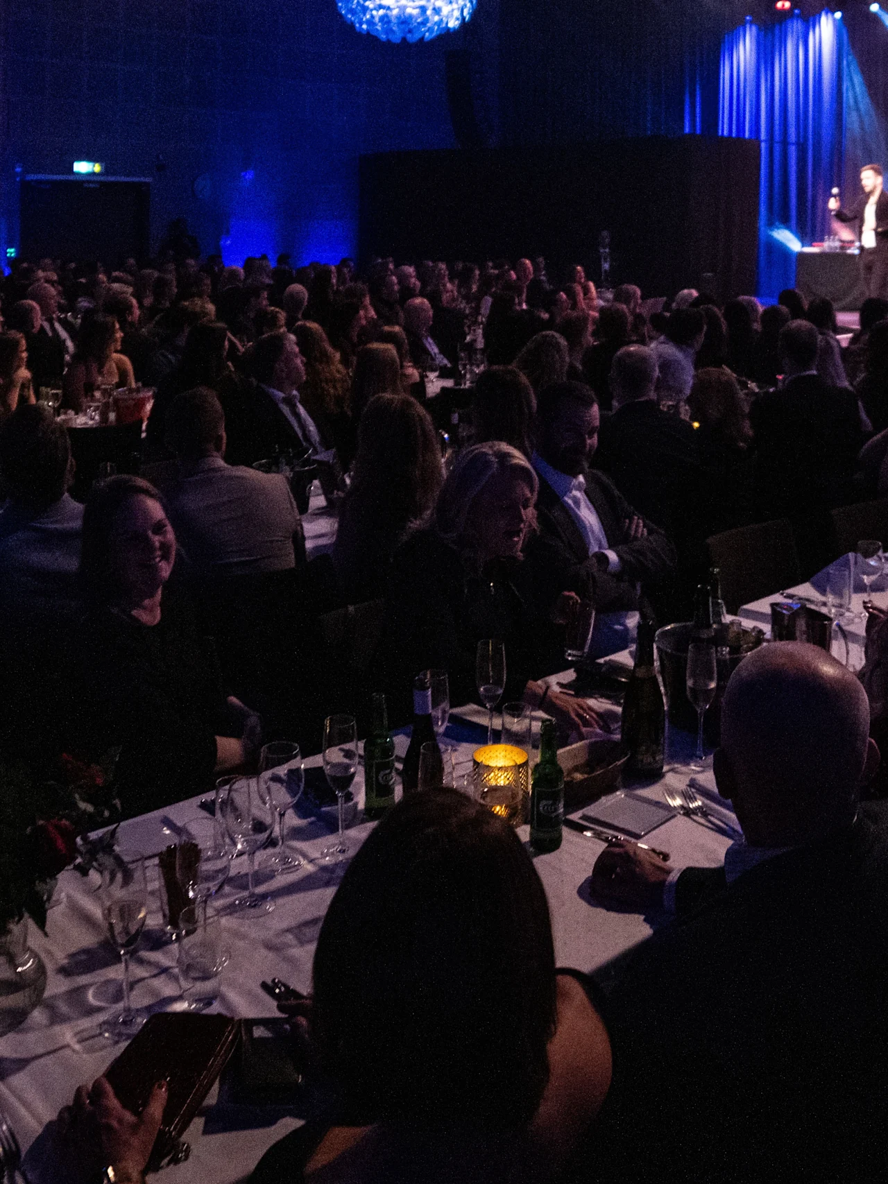 English: A large audience sits at tables, watching a stage performance in a dimly lit room with blue lighting, decorated with chandeliers, creating an elegant atmosphere.