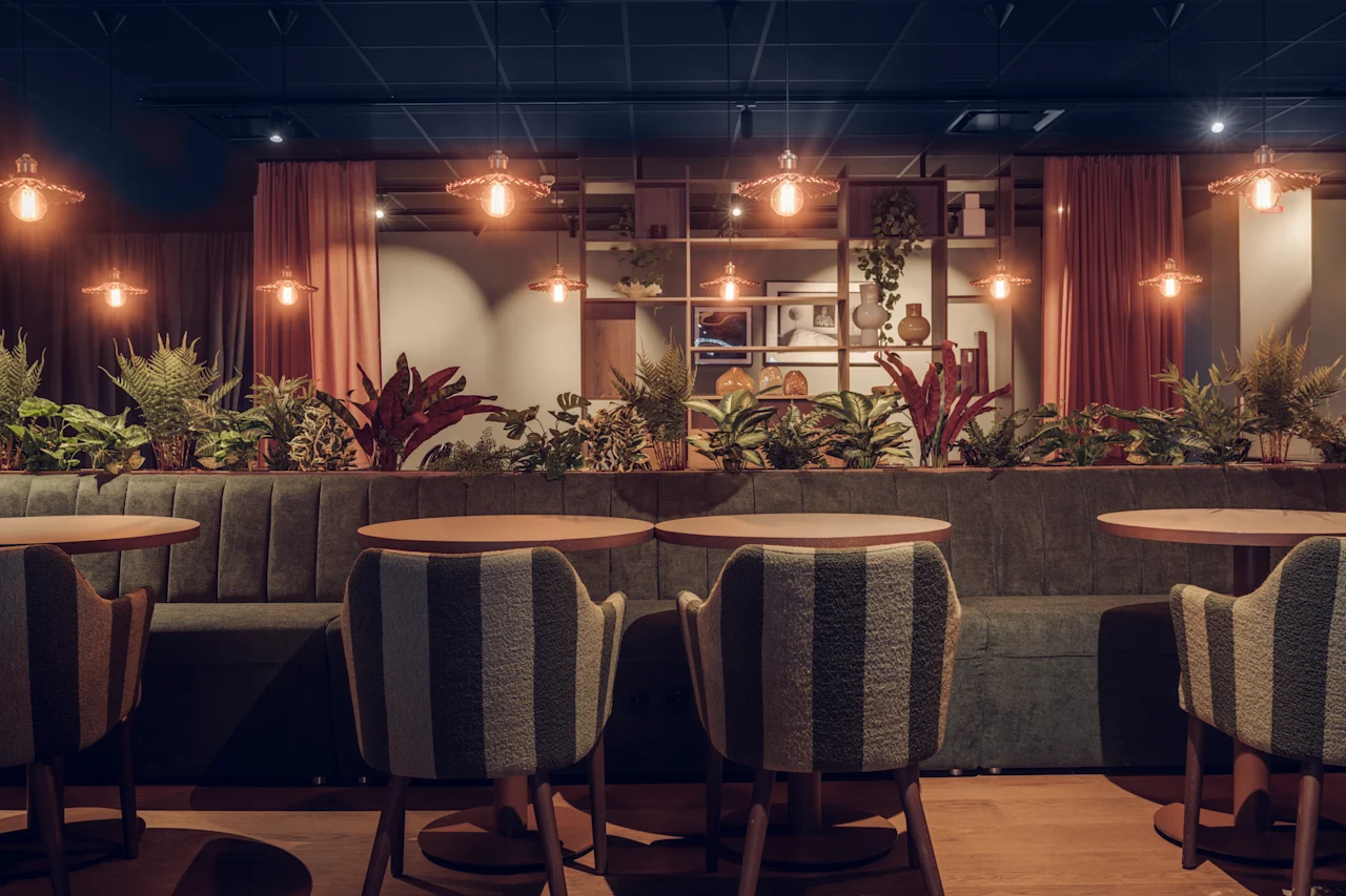 Cozy café setting, soft chairs facing round tables, surrounded by lush plants. Warm pendant lights hang from the ceiling, casting a gentle glow. Shelves with decorative items in the background.