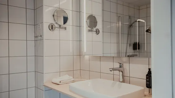 Ett väl upplyst hotellbadrum med ett modernt handfat, stor spegel och en glimt av duschen.