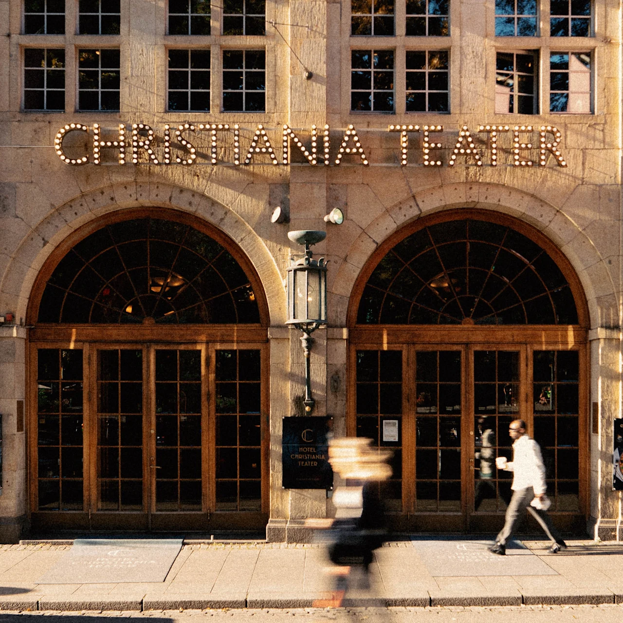 Historic theater facade with "CHRISTIANIA TEATER" in large lighted letters above arched wooden doors, blurred pedestrian and scooter in motion along sidewalk.