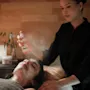 En kvinna får ansiktsbehandling liggandes; en terapeut sprayar ansiktsmist i ett spa. I bakgrunden syns en hylla med hudvårdsprodukter.