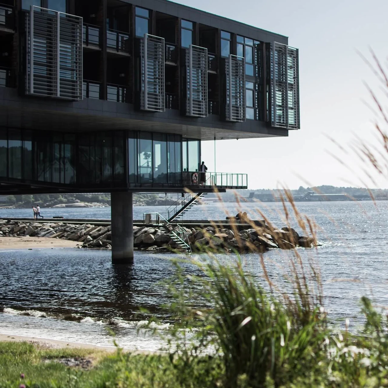 En moderne bygning på pæle over vand, med en person på balkonen og en anden ved strandbredden, foran industrielle strukturer.