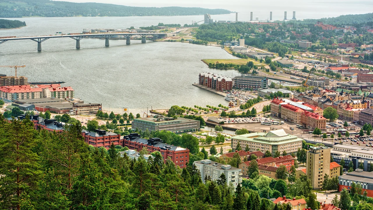Cityscape with buildings stretching along a river, surrounded by lush greenery and a bridge on the left, connecting different parts of the town. Hills and forests frame the scene.